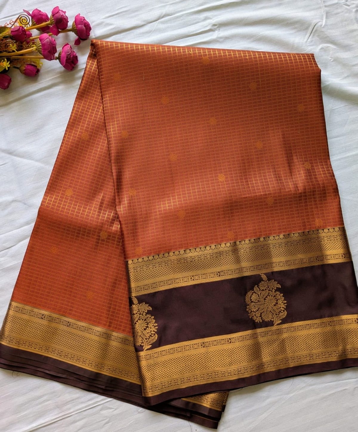 An orange and brown silk saree with gold zari patterns, laid flat on a white fabric background with a small decorative cluster of pink flowers in the top-left corner.